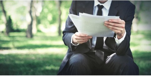 Man dressed in a suit, sitting on a park bench reading papers.