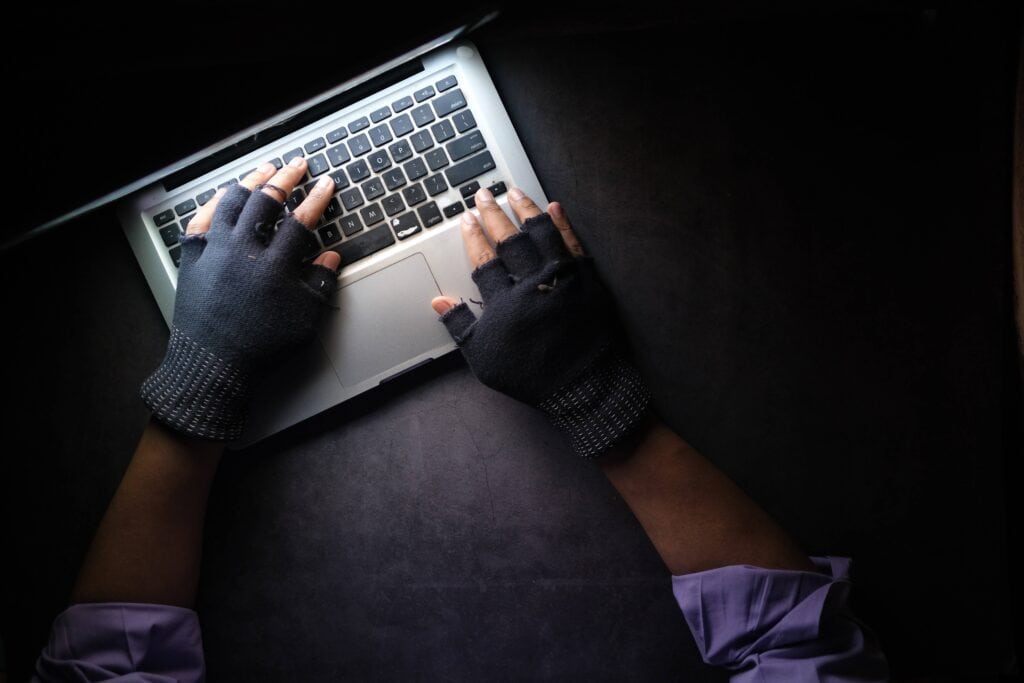 computer sitting on desk with gloved hands typing on it in a dark room.